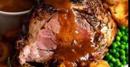 Gigot d’agneau rôti tranché, nappé de sauce au vin blanc et parsemé d’herbes fraîches dans un grand plat de service familial.